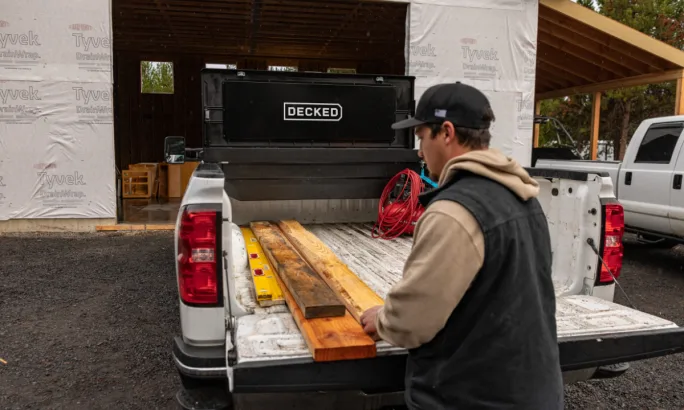 Worker stands at the tailgate with lumber in the bed; a DECKED tool box is mounted at the front of the truck bed.