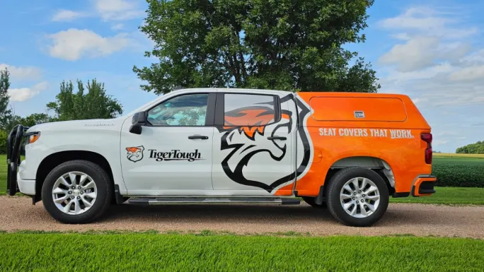 Tiger Tough branded pickup truck with orange and white wrap and tiger logo, parked outdoors, displaying the slogan “Seat Covers That Work.”