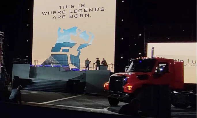Three men on a stage at a distance and a large screen with logo for Mack Trucks and one truck parked in front at the right.
