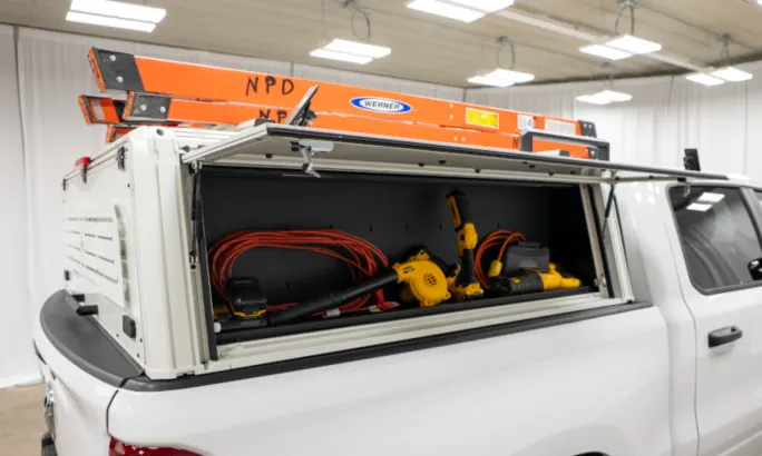 White truck cap cover installed on a white pickup truck and shown with ladders on top and the side open to reveal tool storage.