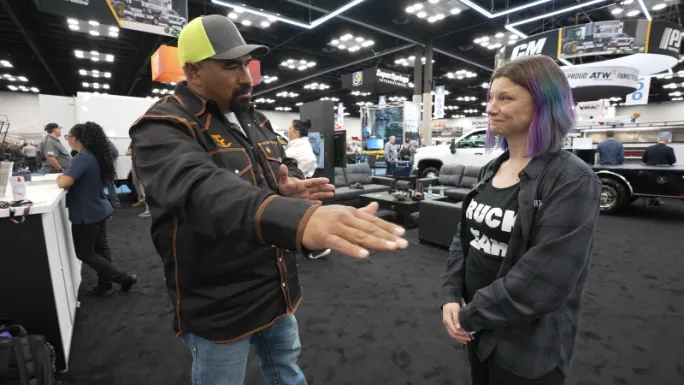 Juan Ibarra gestures while explaining truck clearance challenges to Lauren Fletcher during an interview on a busy Work Truck Week show floor.