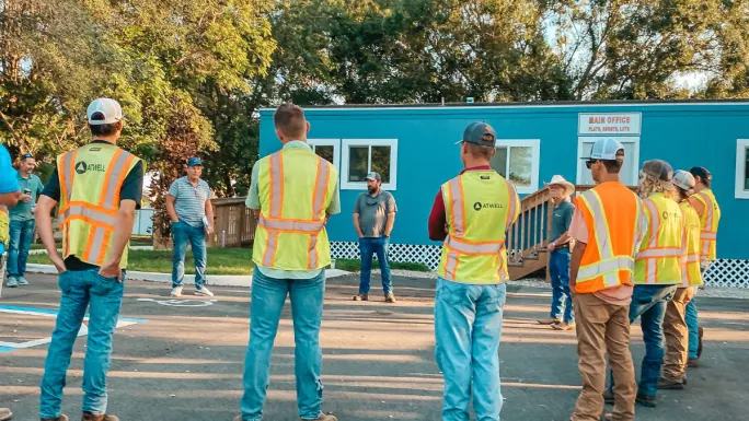 Atwell field employees stand in a circle during an outdoor safety or team meeting, wearing high-visibility vests near a jobsite trailer.