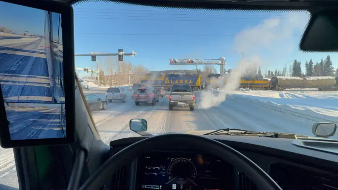 View from inside a truck cab stopped at a snowy Alaska rail crossing, showing traffic, extreme cold conditions, and real-world winter testing environment.