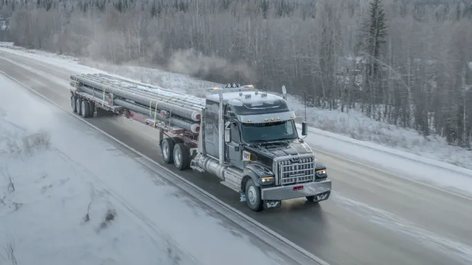 Aerial view of a semi truck hauling pipes on an icy road in winter, demonstrating cold-weather endurance and durability testing in real-world conditions.