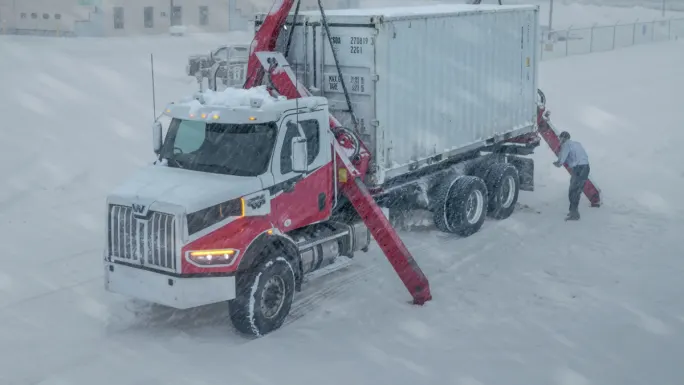 Truck with mounted crane unloading a container in heavy snow, showing equipment performance and reliability during extreme winter operations.