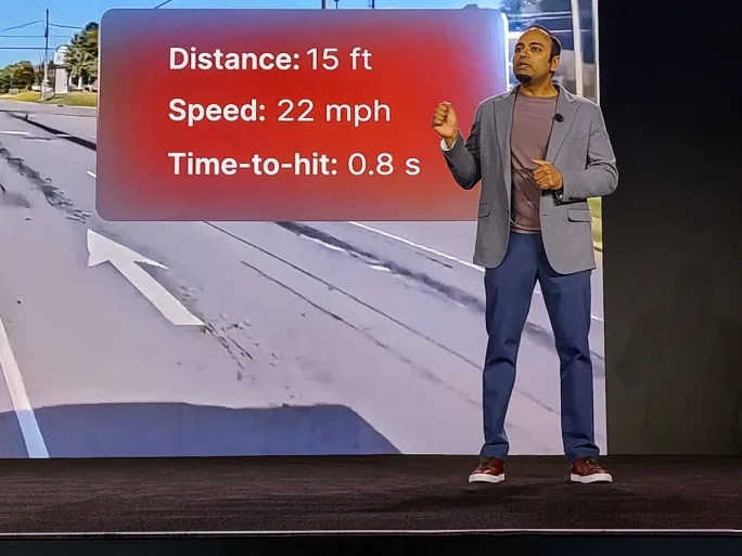 Presenter onstage beside a screen displaying collision alert data: “Distance: 15 ft,” “Speed: 22 mph,” and “Time-to-hit: 0.8 s,” over a roadway background.