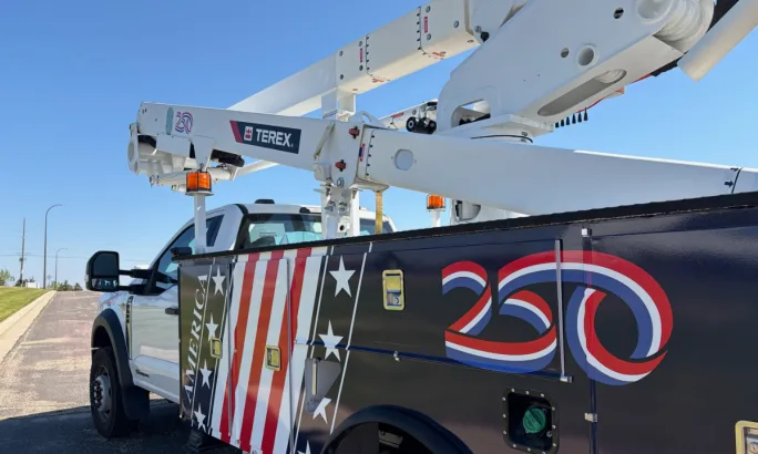 White utility service truck with a Terex aerial lift parked outdoors. The truck body features an American flag-themed graphic and a large red, white, and blue “250” emblem, commemorating a milestone or anniversary.