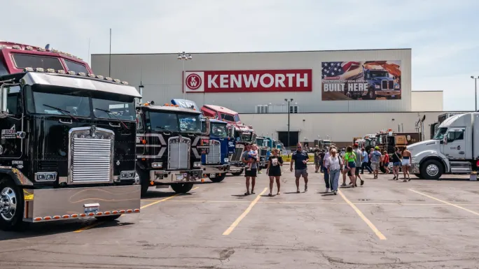 Crowds walk among rows of customized Kenworth trucks parked outside a Kenworth manufacturing facility. The event features show trucks, attendees, and a large Kenworth sign on the building in the background.