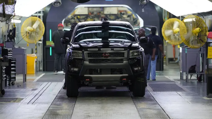 Front view of a black Nissan Frontier pickup truck on the assembly line at Nissan's Canton manufacturing facility with employees working nearby.