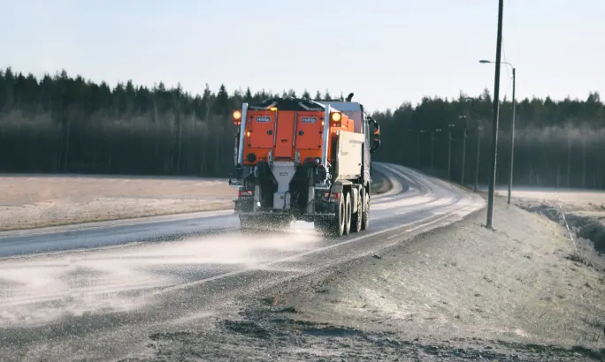 A road maintenance truck applies de-icing material to a two-lane rural roadway, spraying the pavement as it travels through a lightly frosted landscape bordered by fields and trees.