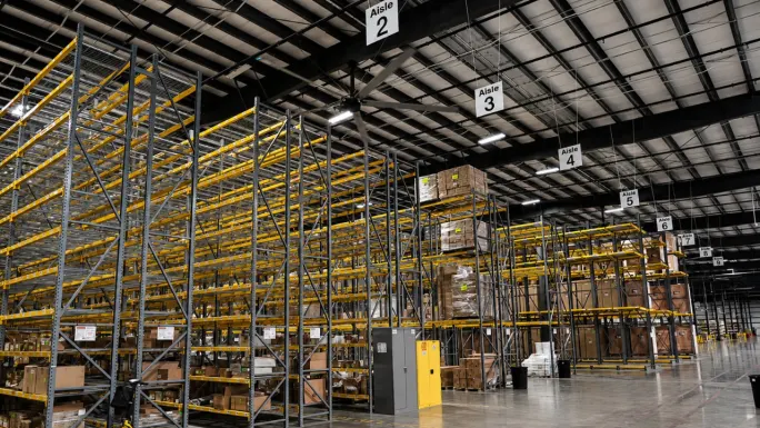 Interior of a large BendPak warehouse with tall pallet racking, labeled storage aisles, and boxed inventory organized on multiple levels beneath a high industrial ceiling, highlighting extensive parts storage and distribution capabilities.