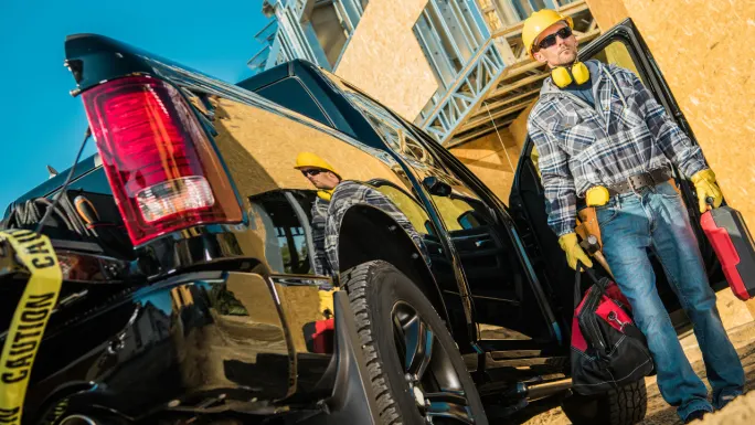 Construction worker in a hard hat stands beside an open black pickup at a jobsite, carrying tool cases and a work bag.