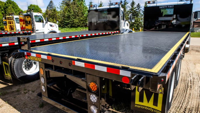 Black Marion platform truck body with an open flat deck and safety bulkhead, photographed outdoors with additional work trucks in the background.