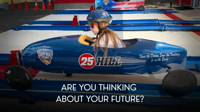 A young racer wearing a helmet sits inside a blue Soap Box Derby car at the starting area beneath the words “Are You Thinking About Your Future?”