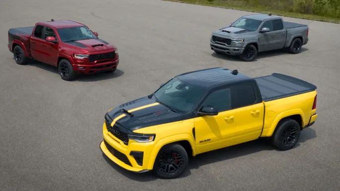 Three 2027 Ram 1500 Rumble Bee muscle trucks in yellow, red, and gray parked on pavement.