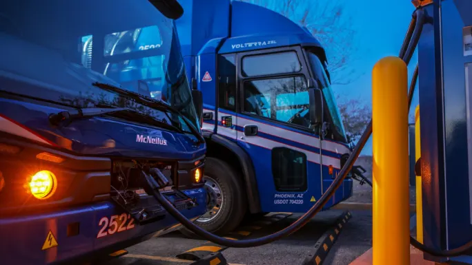 Two blue Republic Services electric refuse trucks parked at a charging station, with one truck connected to a charger by a heavy-duty cable.