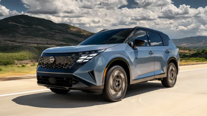 Blue 2027 Nissan Rogue Hybrid driving on a mountain road under a partly cloudy sky.