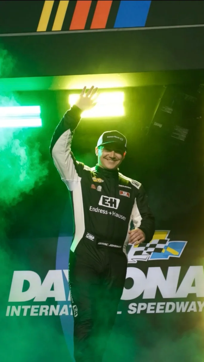 Isaac Johnson in a black-and-white firesuit waves under bright green and yellow lights during a driver introduction at Daytona International Speedway.