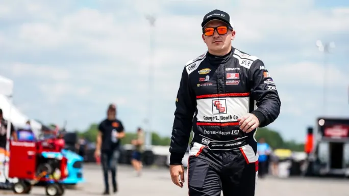 Isaac Johnson wearing a black, white, and red firesuit, cap, and orange sunglasses walks through a busy motorsports paddock.
