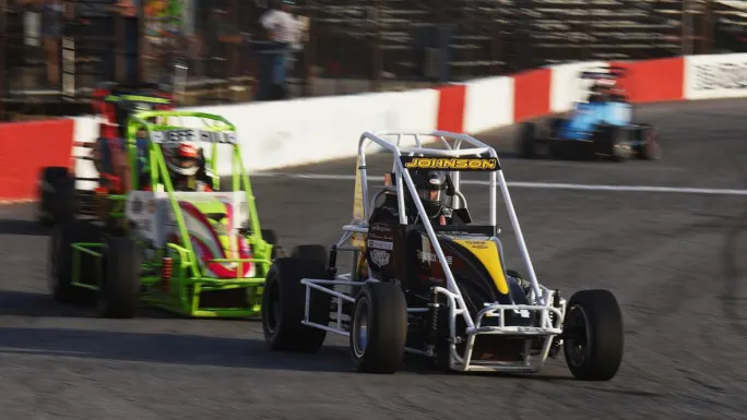 Two open-wheel race cars compete on a paved short track, with a black-and-yellow car marked Johnson leading a green car through a turn.