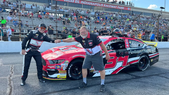 Isaac Johnson and a team member bump fists beside a red, black, and white No. 34 race car at Lucas Oil Indianapolis Raceway Park as spectators watch from the grandstands.