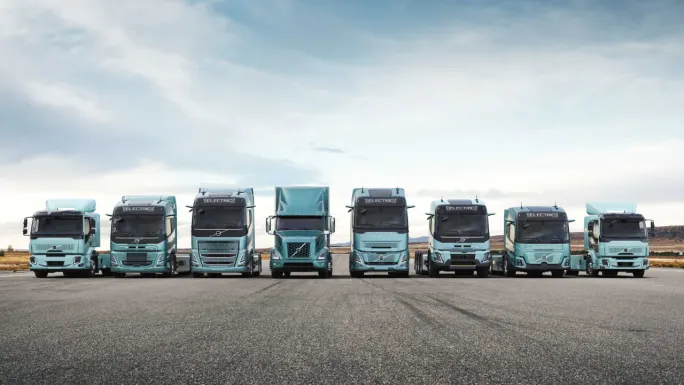 Lineup of eight Volvo electric commercial trucks in various configurations parked side by side on an open roadway beneath a cloudy sky.