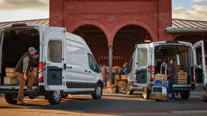 Two Ford Transit Cargo Vans with rear doors open as workers load produce outside a market.