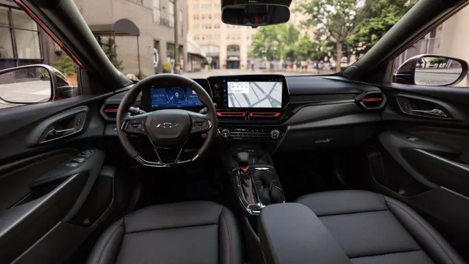 Wide interior view of the 2027 Chevrolet Trax showing the steering wheel, digital instrument cluster, center touchscreen displaying navigation, front seats, and center console with red dashboard accents. Preproduction model shown.