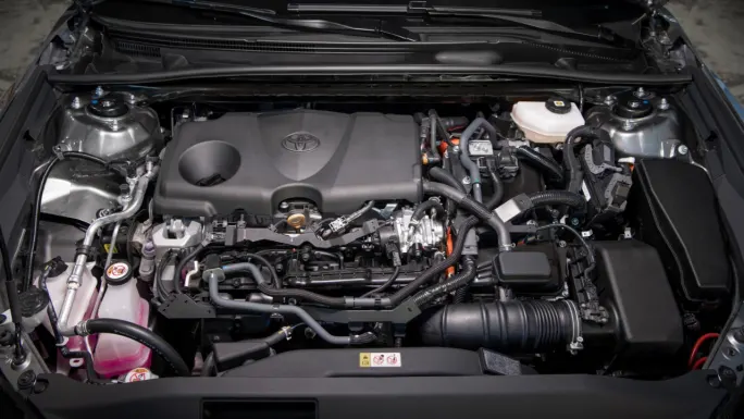 Overhead view of the 2027 Toyota Camry's engine compartment, showing the Toyota-branded engine cover, hoses, wiring, fluid reservoirs, and other components of the hybrid powertrain.