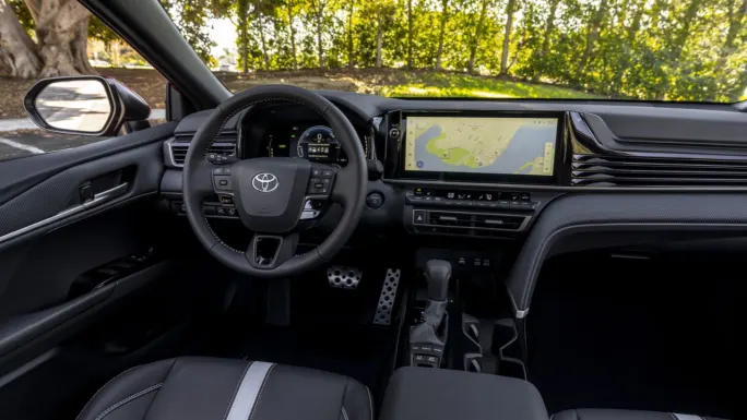 Driver's view of the 2027 Toyota Camry interior, featuring a Toyota-branded steering wheel, digital instrument cluster, large center touchscreen displaying navigation, and center console.