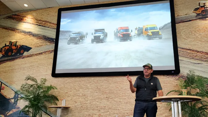 A presenter speaks beside a large screen displaying Volvo vocational trucks driving through a dusty quarry, with construction equipment imagery decorating the walls of the presentation room.