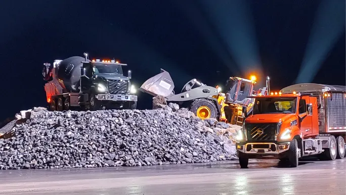 Volvo VNS vocational trucks operating at a nighttime construction site, including a concrete mixer, wheel loader, and orange tractor-trailer beside a large pile of rocks, illuminated by vehicle headlights and work lights.