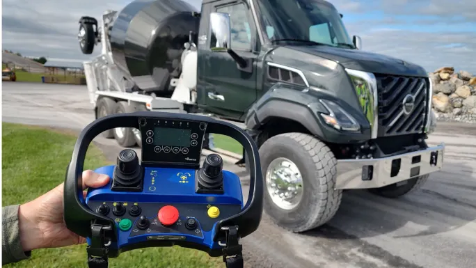 Close-up of a handheld remote steering controller with dual joysticks and control buttons, held in front of a dark green Volvo VNX concrete mixer truck at an outdoor worksite.