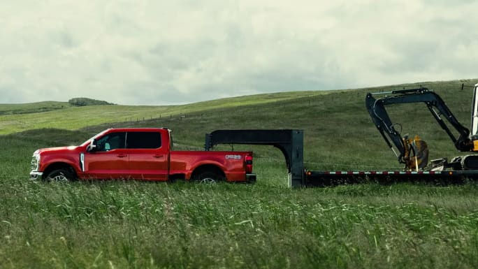 Red Ford Super Duty Crew Cab towing a gooseneck flatbed trailer carrying an excavator through a grassy field.