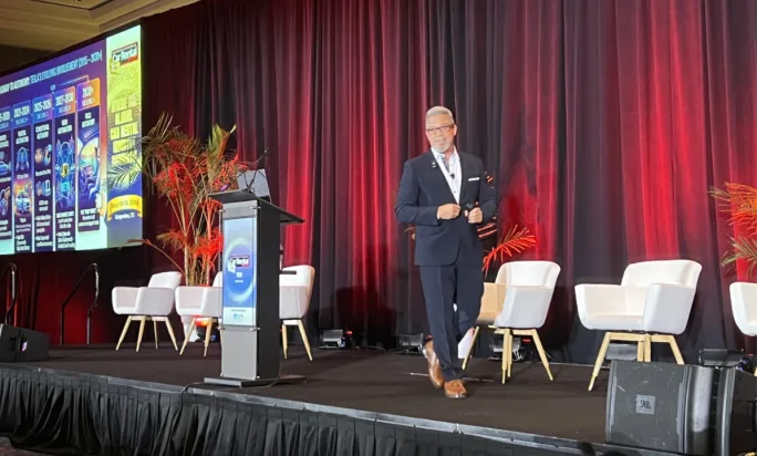 Keynote speaker on a stage in front of a burgundy curtain and large video screen with an autonomous mobility display.