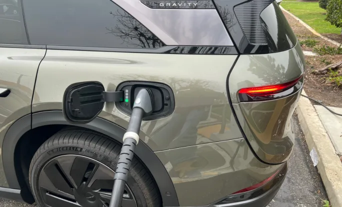 A green-gray Lucid Gravity SUV being charged at left rear charging port at a parking lot charging station.