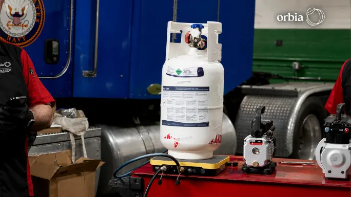 A cylinder labeled R-456A sits on a refrigerant scale beside service equipment, with a blue truck in the background.
