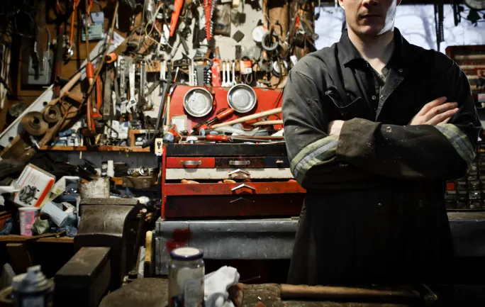 Mechanic in a cluttered workshop stands with arms crossed beside a red toolbox, symbolizing skilled technicians prepared for emergency equipment repairs.