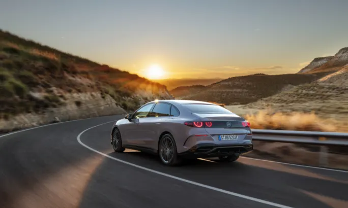 Rear view of a silver Mercedes-Benz electric sedan driving on a mountain road at sunset with glowing taillights.