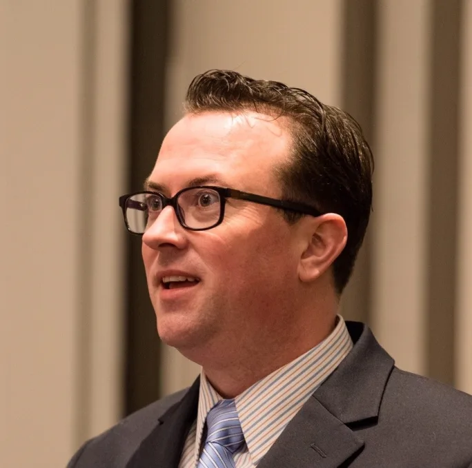 A portrait of a man in a suit and glasses looking off camera left.