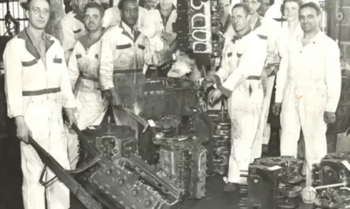 A black and white photo shows employees posing with vehicle transmissions.