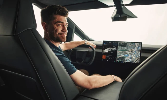 A bearded young man at the steering wheel of a Tesla Cybertruck, looking over high right shoulder toward the rear seat.