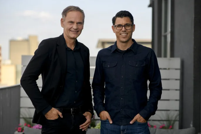 Two men pose in on an outdoor balcony in dark colored business casual attire.