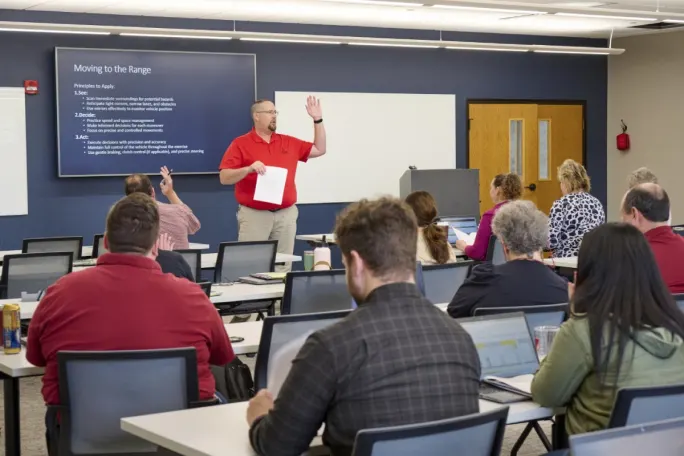 Adult classroom setting with a teacher standing in front of a projector screen addressing the class.