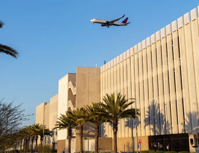 A passenger jet flies over the LAX CONRAC building, lined with palm trees.