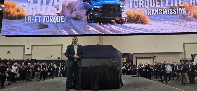 Ram executive Matt Huber stands on stage speaking to an audience at Work Truck Week while a covered vehicle and large Ram display screen appear behind him during the ProMaster City presentation.