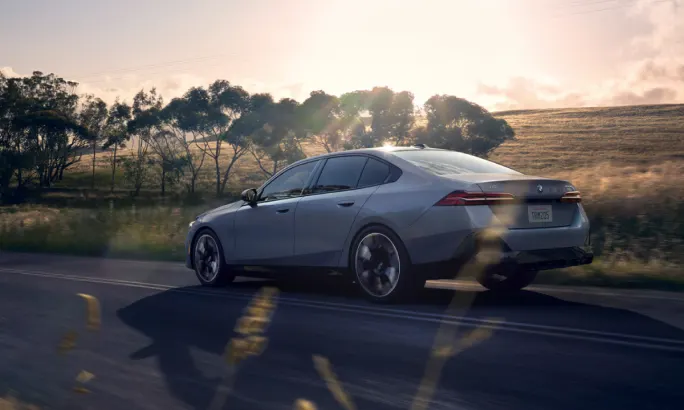 The left side of a gray 2025 BMW i5 series vehicle driving on a road surrounding by fields and trees.