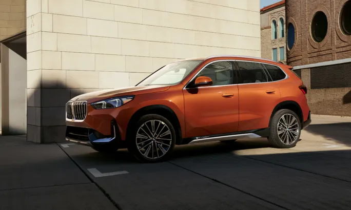 An orange 2025 BMW X1 model parked in by the side of a sunlit gray brick building.