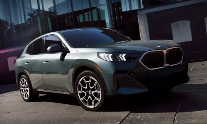 An up close front angle shot of a blue green 2025 BMW X2 in front of a glass building.
