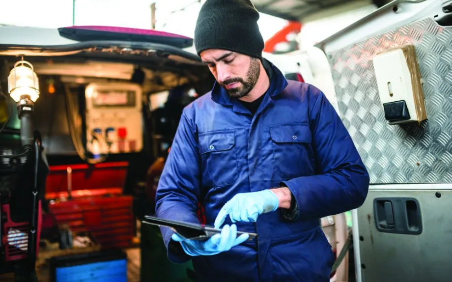 Fleet technician wearing gloves and a blue work jacket reviews information on a tablet inside a mobile service vehicle.